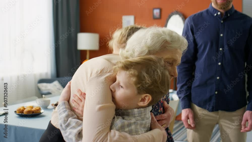 Joyous elderly grandmother giving hugs to little grandkids while greeting family at home for holiday dinner Joyous elderly grandmother giving hugs to little grandkids while greeting family at home for holiday dinner