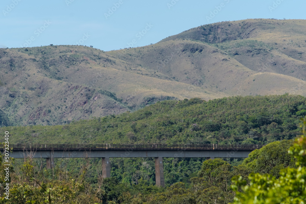Vista de longe, linha ferrea construída sobre ponte aos pés de montanha, cercado de muita vegetação e lindo céu azul, na região do Parque das águas, localizado no Barreiro, Belo horizonte, Minas Gerai