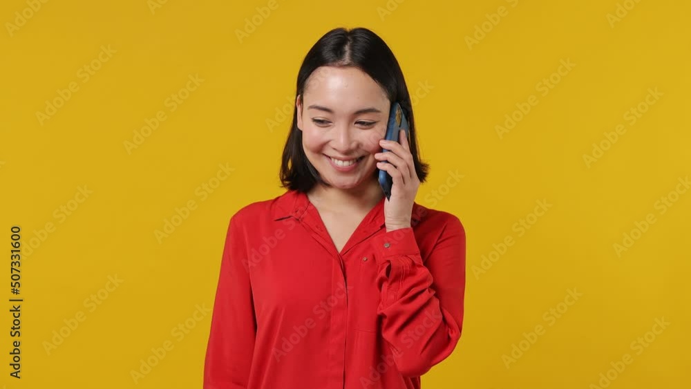 Happy calm smiling young woman of Asian ethnicity 20s years old wears red shirt hold use talk on mobile cell phone conducting pleasant conversation isolated on plain yellow background studio portrait