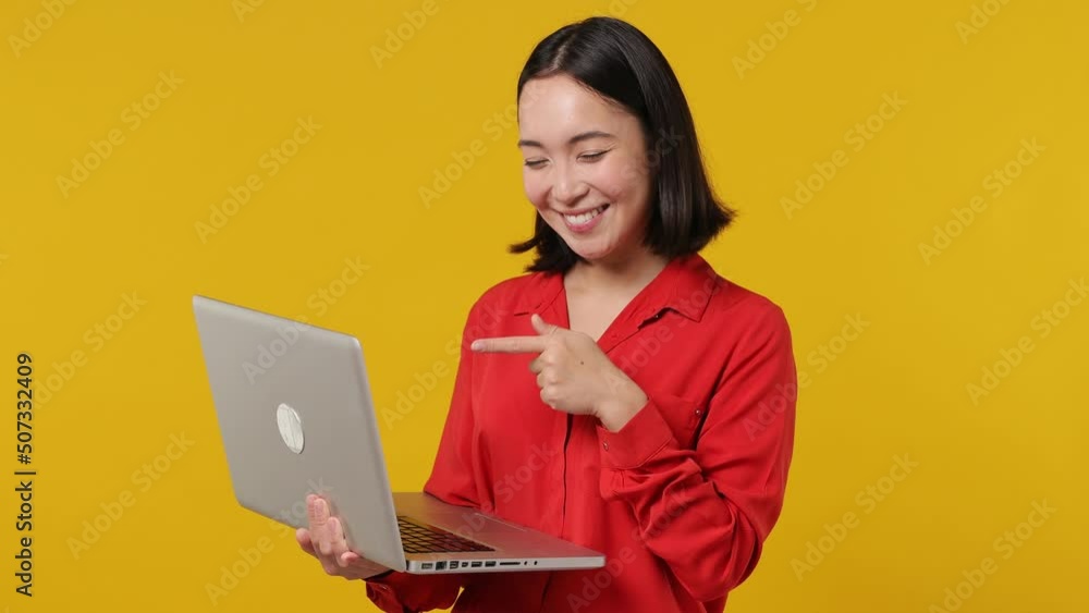 Smiling businesslike young woman of Asian ethnicity 20s years old wears red shirt hold use work point finger on laptop pc computer typing chatting isolated on plain yellow background studio portrait Smiling businesslike young woman of Asian ethnicity 20s years old wears red shirt hold use work point finger on laptop pc computer typing chatting isolated on plain yellow background studio portrait