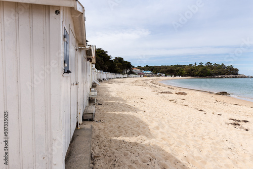 Baie sableuse avec cabines de plage blanches alignées et forêt en arrière-plan. Plage des Dames à Noirmoutier