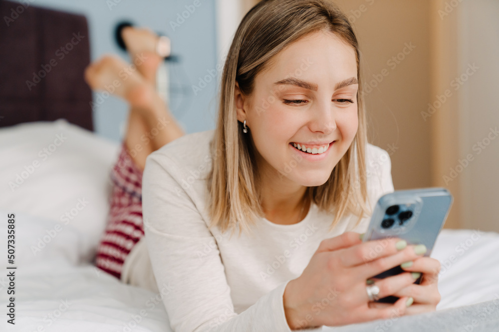 Young white blond woman smiling and using cellphone in bed at home Young white blond woman smiling and using cellphone in bed at home