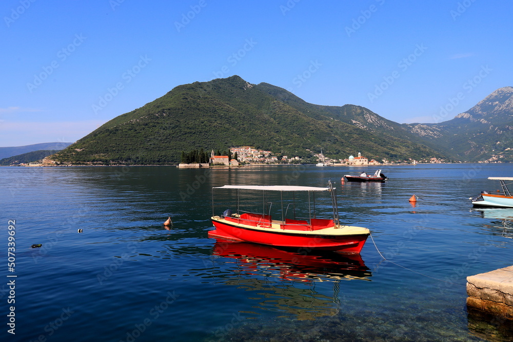 Boka Kotor, Montenegro. A red boat sails along Bay Kotor against backdrop of famous island and Church Madonna on Reef, Perast