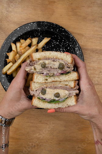 Top view of caucasian female hands holding a a Vitel tonné sandwich with veal, cream, capers, arugula and french fries. photo