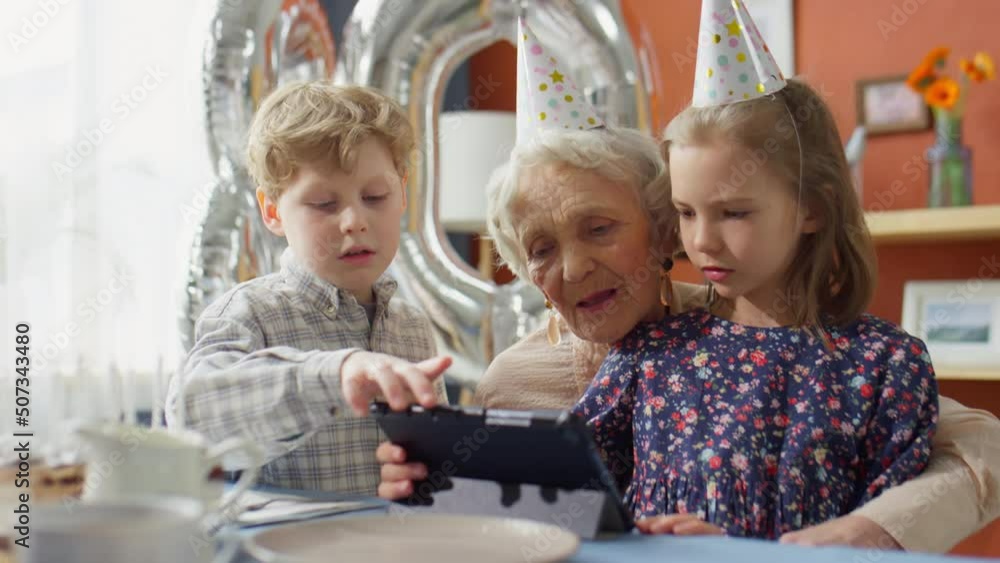 Little boy and girl teaching elderly grandmother how to use digital tablet while having birthday dinner party at home Little boy and girl teaching elderly grandmother how to use digital tablet while having birthday dinner party at home
