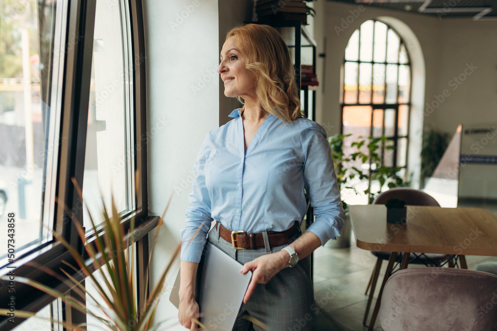 Mature businesswoman holding laptop standing in cafe seen from window. Mature businesswoman holding laptop standing in cafe seen from window.