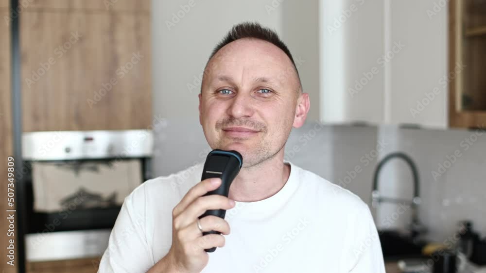 funny handsome caucasian man guy male in white t shirt shaving with electric razor having fun shaving in kitchen at home dancing music humorous hygiene. funny handsome caucasian man guy male in white t shirt shaving with electric razor having fun shaving in kitchen at home dancing music humorous hygiene.