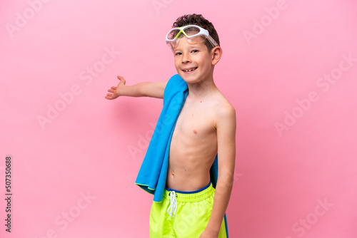 Little caucasian boy wearing a diving goggles isolated on pink background extending hands to the side for inviting to come