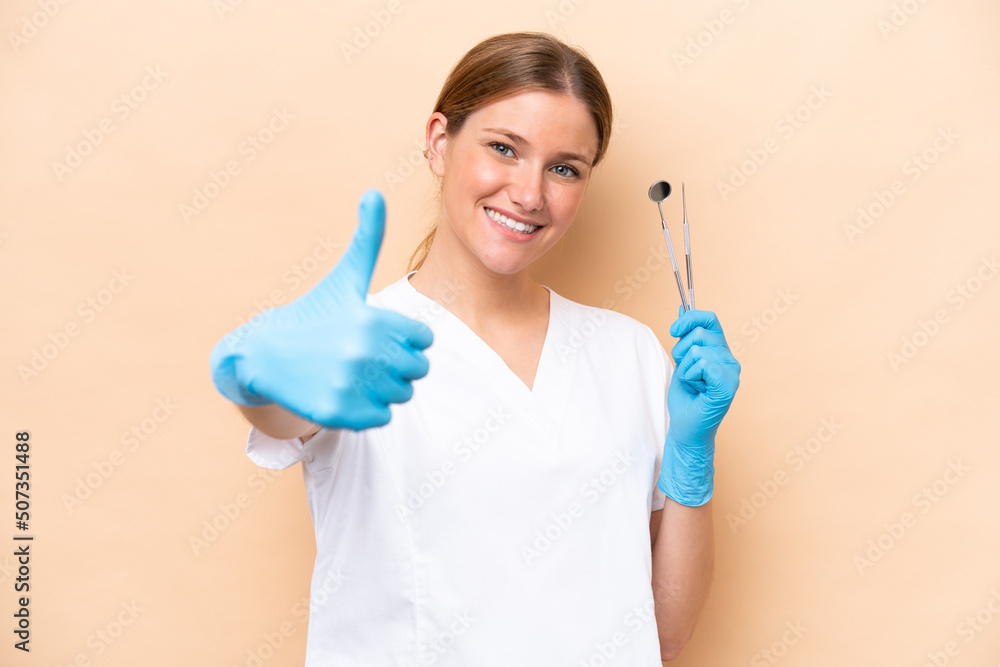 Dentist caucasian woman holding tools isolated on beige background with thumbs up because something good has happened