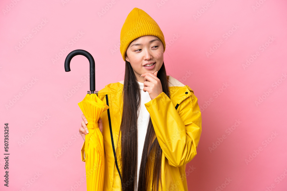 Young Chinese woman with rainproof coat and umbrella isolated on pink background looking to the side and smiling Young Chinese woman with rainproof coat and umbrella isolated on pink background looking to the side and smiling
