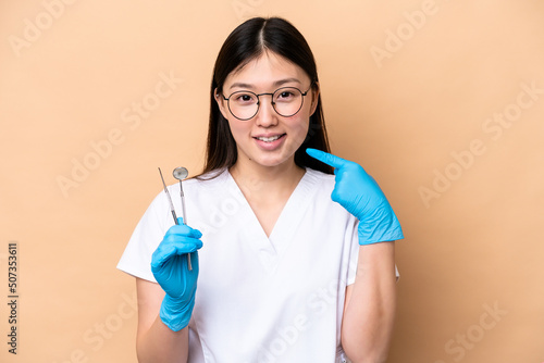 Dentist Chinese woman holding tools isolated on beige background giving a thumbs up gesture