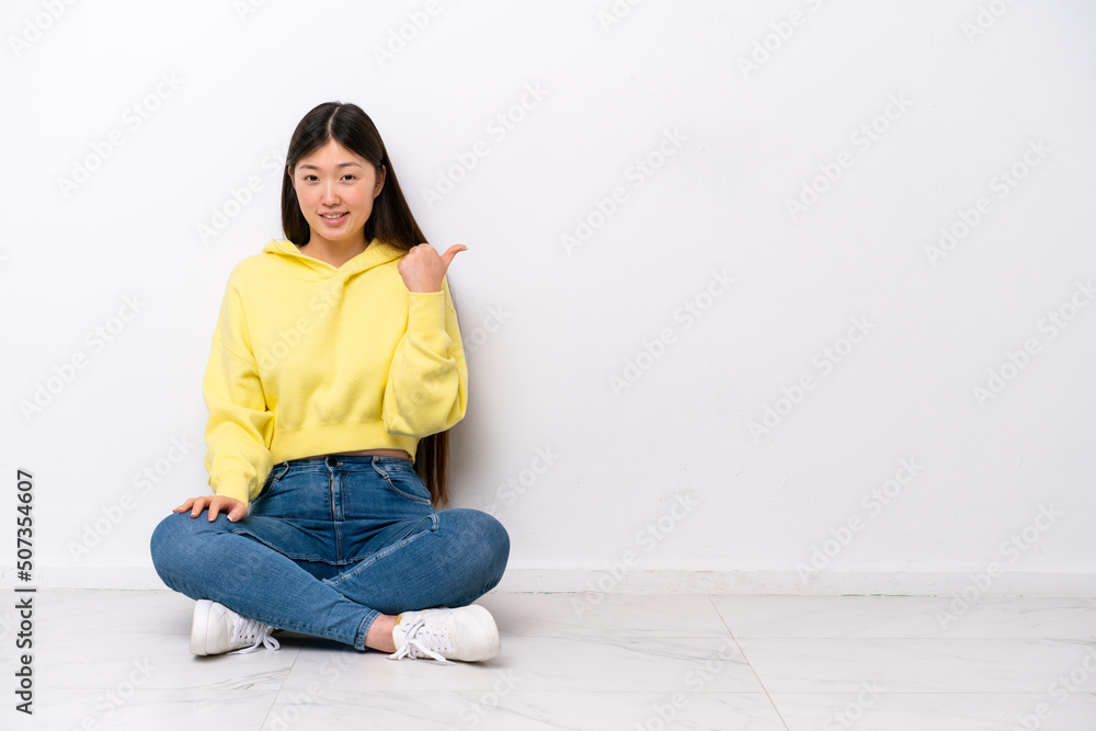Young Chinese woman sitting on the floor isolated on white wall pointing to the side to present a product Young Chinese woman sitting on the floor isolated on white wall pointing to the side to present a product