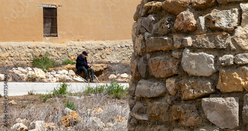 Photos an old man in a beret sits in front of a stone wall in a small, uninhabited Span