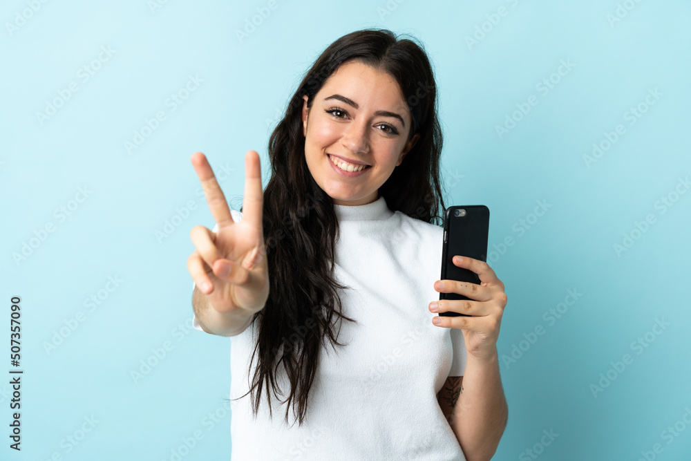 Young woman using mobile phone isolated on blue background smiling and showing victory sign Young woman using mobile phone isolated on blue background smiling and showing victory sign