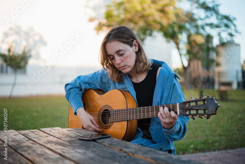 Girl enjoying with her guitar while reading music sheet on her phone. She is sitting in a wooden picnic area.