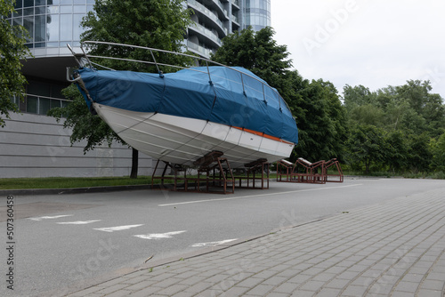 A motor boat covered with a blue cover stands on land under the open sky against the background of trees