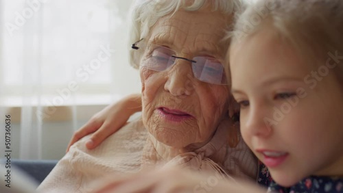 Close up shot of little girl reading book together with elderly grandmother at home