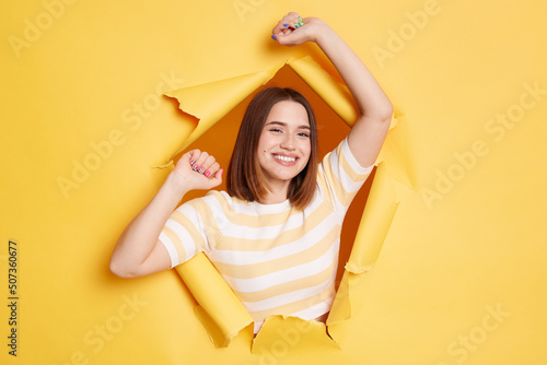 Horizontal shot of attractive beautiful woman stands in torn paper hole, posing with raised arms and looking at camera with charming smile, looking through breakthrough of yellow background. © sementsova321