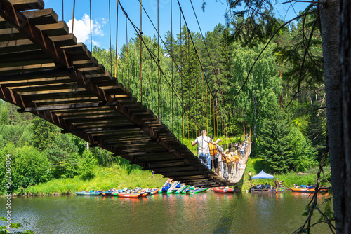 People in life jackets cross the suspension bridge over the Chusovaya River. Parking of colorful boats and catamarans for rafting. Active lifestyle and sports