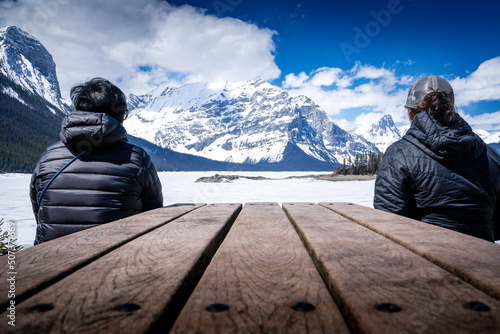 Two people in nature sit at a table overlooking a frozen lake with snow covered mountains in the Canadian Rockies at Upper Kananaskis Lake Alberta Canada. photo