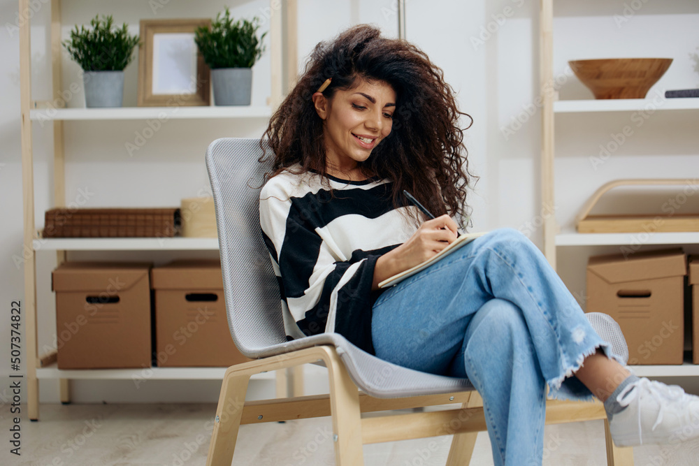 Happy concentrated pretty beautiful curly tanned Latin woman in striped shirt hold notebook look down write work letter essay in home interior. Copy space Banner. Writer concept Happy concentrated pretty beautiful curly tanned Latin woman in striped shirt hold notebook look down write work letter essay in home interior. Copy space Banner. Writer concept