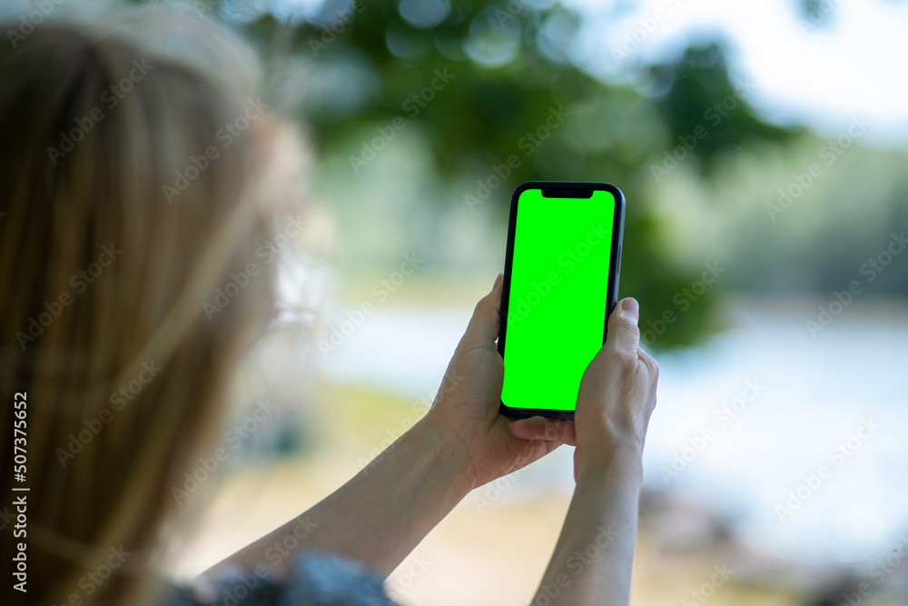 Blonde woman holding phone with empty greenscreen and taking pictures of lake. Mobile photography