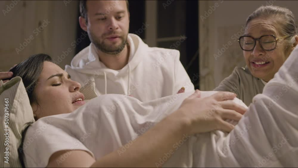 Pregnant woman lying on bed and having contractions at home. Caucasian woman suffering from pain. Asian doula and husband stroking belly and talking with expecting mother. Medicine, pregnancy concept Pregnant woman lying on bed and having contractions at home. Caucasian woman suffering from pain. Asian doula and husband stroking belly and talking with expecting mother. Medicine, pregnancy concept