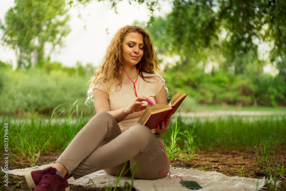 A beautiful young curly hair woman sitting on blanket in grass while reading a book and listening music. A beautiful young curly hair woman sitting on blanket in grass while reading a book and listening music.