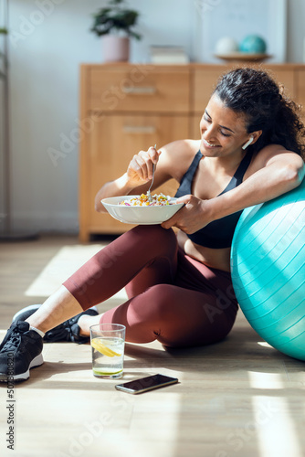 Sporty young woman eating healthy while listening to music sitting on the floor at home.