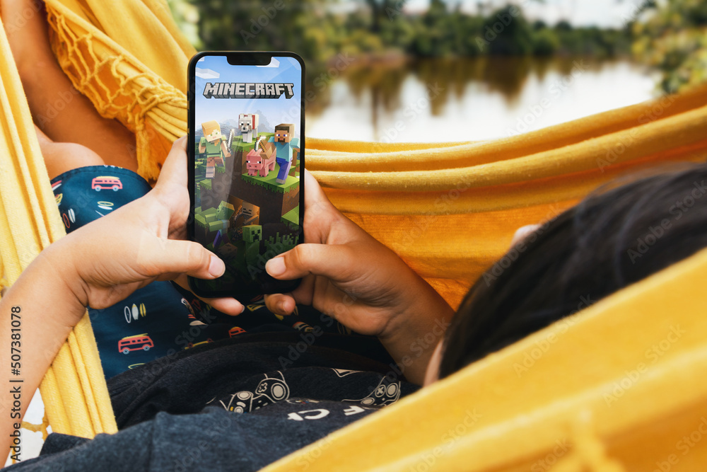 Naklejka premium Child lying in hammock with Minecraft mobile game app on smartphone screen. Lake in the background. Rio de Janeiro, RJ, Brazil. May 2022