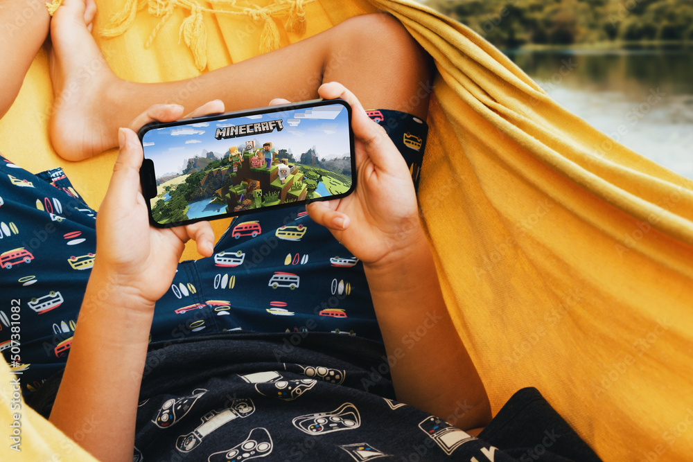 Naklejka premium Child lying in hammock with Minecraft mobile game app on smartphone screen. Lake in the background. Rio de Janeiro, RJ, Brazil. May 2022