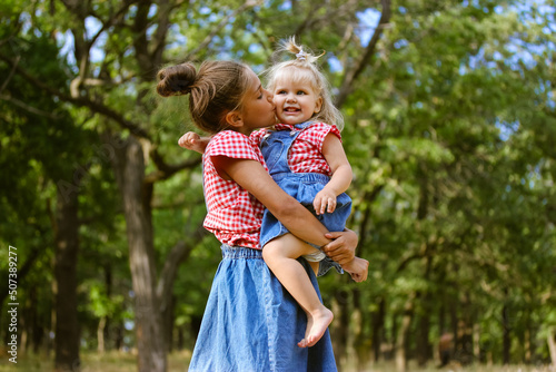 Cute sisters happy together celebrating Children's Day June 1 outdoors. Older sister hugging kissing baby girl. Kids activities in summer camp. Cousins have fun in park, forest. Little girls on nature