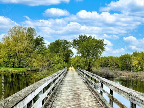 Under a sunny blue sky on a summer day the Great River State Trail crosses a Mississippi River backwater on a wooden trestle passing through lush green forests reflected in the still waters.