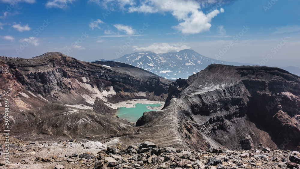 Obraz premium Amazing acid turquoise lake on top of an active volcano. The layered structure of the steep slopes of the crater, rocky soil is visible. The mountain against the blue sky. Kamchatka. Gorely Volcano