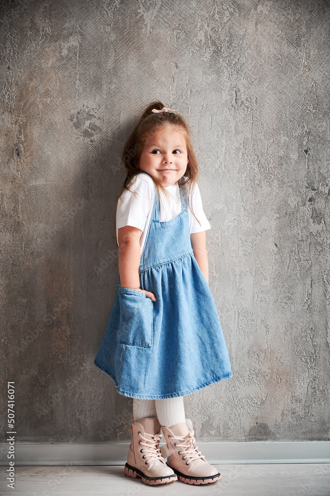 Full length of adorable little girl in denim dress standing against the wall in new home. Cute female child keeping hand in pocket. Full length of adorable little girl in denim dress standing against the wall in new home. Cute female child keeping hand in pocket.