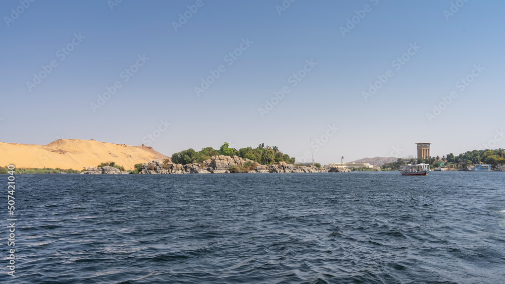 Tourist boats sail on the blue river. Picturesque boulders and green vegetation are visible on the shore. A sand dune against a clear sky. Egypt. Nile. Copy Space