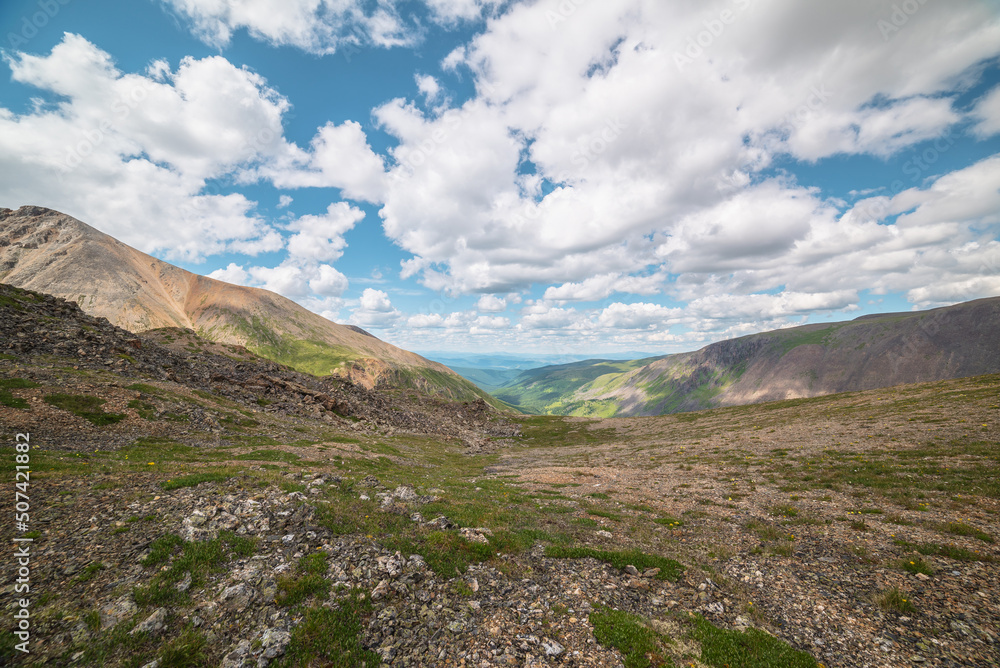 Scenic view from mountain pass to green forest valley among mountain ranges and hills on horizon at changeable weather. Green landscape with sunlit mountain vastness under cumulus clouds in blue sky. Scenic view from mountain pass to green forest valley among mountain ranges and hills on horizon at changeable weather. Green landscape with sunlit mountain vastness under cumulus clouds in blue sky.