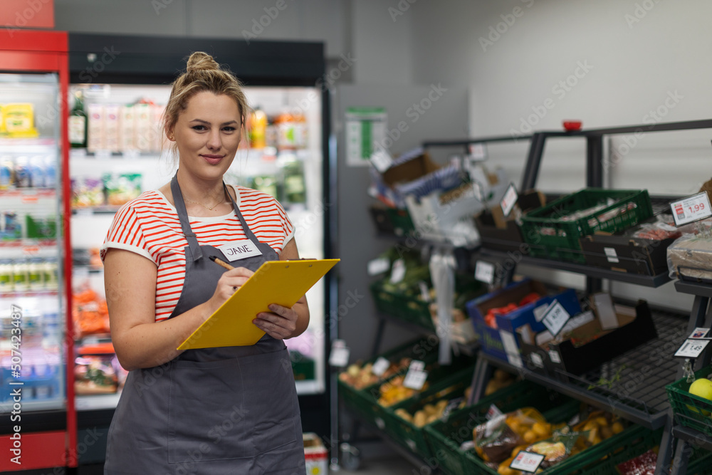 A young shop assistant in supermarket standing and writing in front of vegetable section.