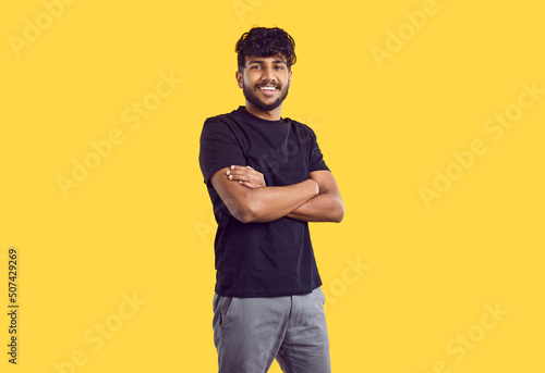 Studio shot of happy young man in casual wear. Handsome Indian guy with modern haircut in black Tshirt standing with his arms folded isolated on yellow background, looking at camera and smiling