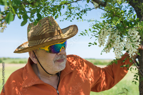 Portrait of bearded Ukrainian senior farmer standing under  sadow of flowering acacia tree against of green agricultural field and looking forward.
