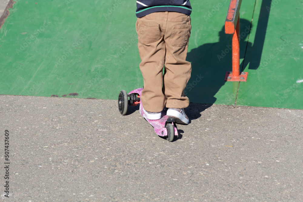 Obraz premium A little boy in brown pants and a cap is having fun and diligently studying extreme sports, riding a scooter in summer on the playground, in the park and outdoors.