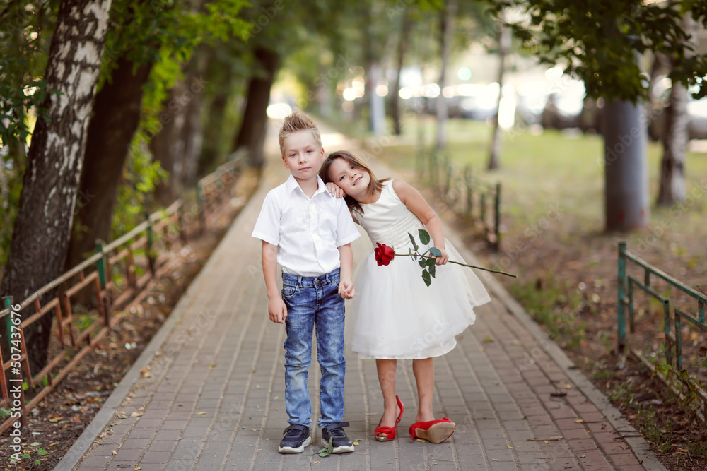 Fototapeta premium Cute couple children boy and girl hugs and holding rose on walk in summer, children play date and courtship, romantic and growing up concept