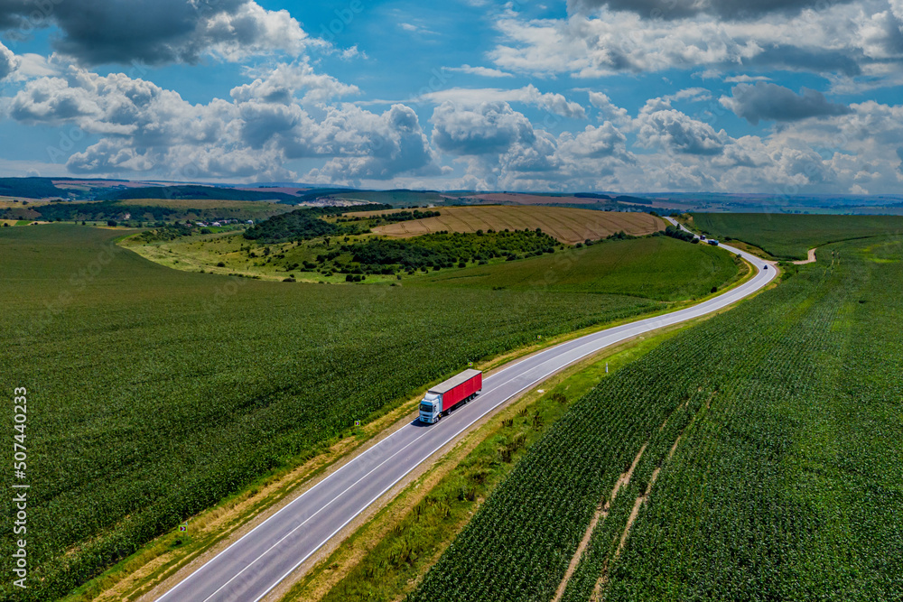 red truck driving on asphalt road along the green fields. beautiful clouds in the sky. Aerial view landscape. drone photography. cargo delivery and transportation concept.