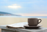 Ceramic cup of hot drink and newspaper on stone surface near sea in morning. Space for text