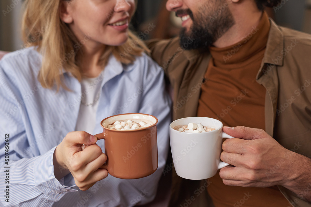 Stylish bearded man and young woman sitting together on sofa at home holding cups of hot coffee with marshmallow, copy space