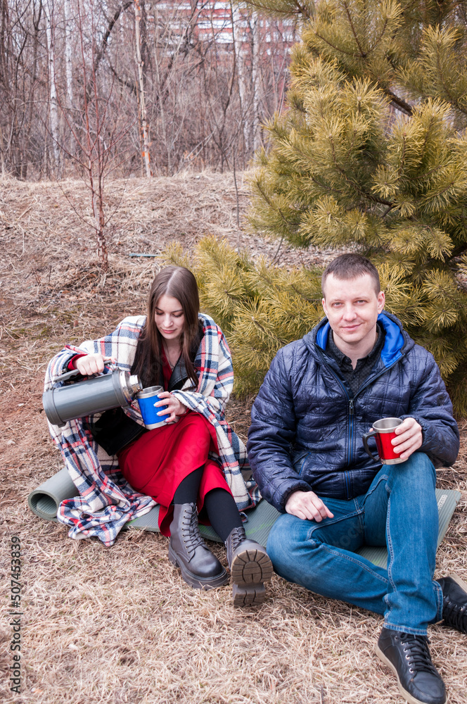 A couple in a spring park: a man and a girl sit near a fir tree and drink hot tea from a thermos