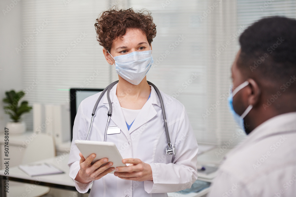 Horizontal over-the-shoulder shot of modern female doctor wearing white coat and protective mask consulting patient, copy space Horizontal over-the-shoulder shot of modern female doctor wearing white coat and protective mask consulting patient, copy space