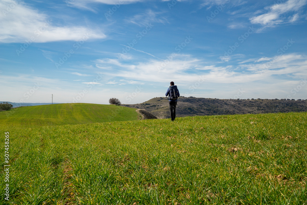 Unrecognizable young man walking with a hiking backpack through a green meadow in the middle of nature Unrecognizable young man walking with a hiking backpack through a green meadow in the middle of nature