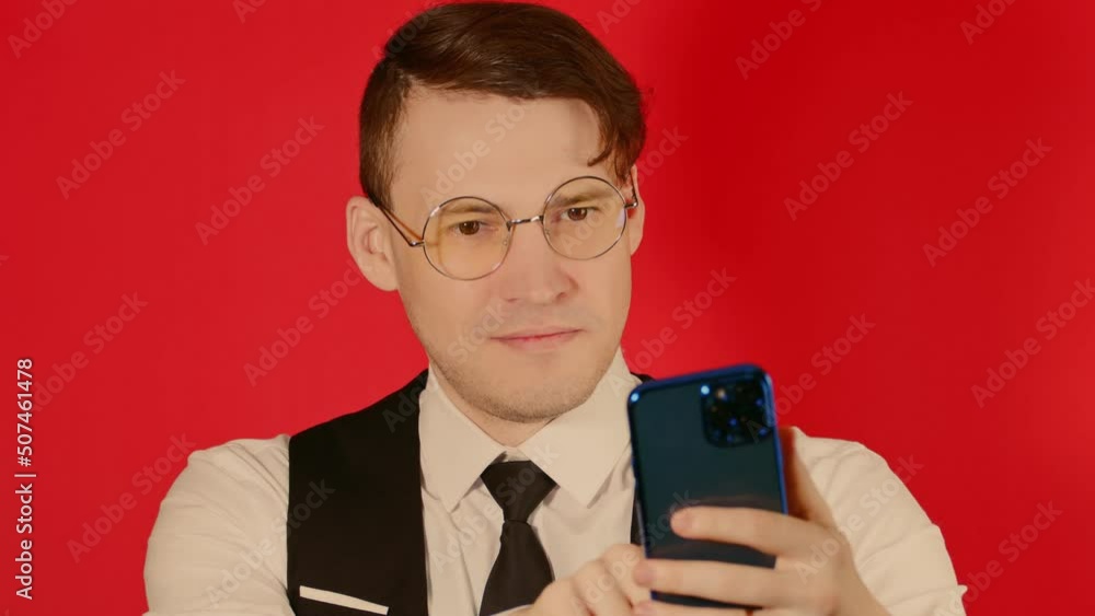 Smiling man using cellphone in studio.Young male in formal suit and glasses using smartphone while standing against red background Smiling man using cellphone in studio.Young male in formal suit and glasses using smartphone while standing against red background