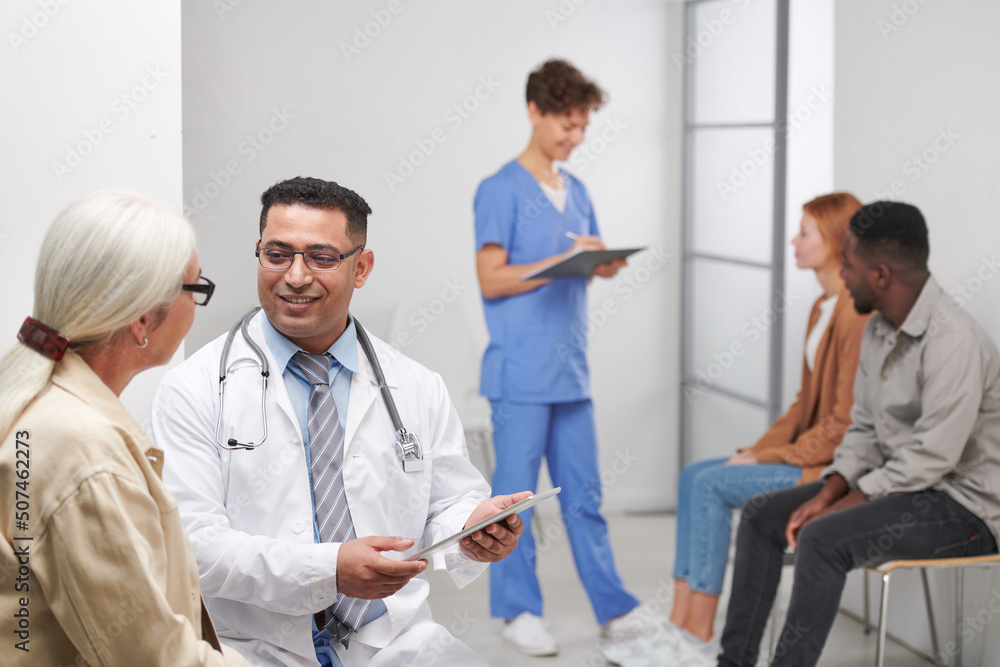 Obraz premium Modern Middle-Eastern medical worker talking to senior woman, his female colleague calling for next patient in queue and making notes on background